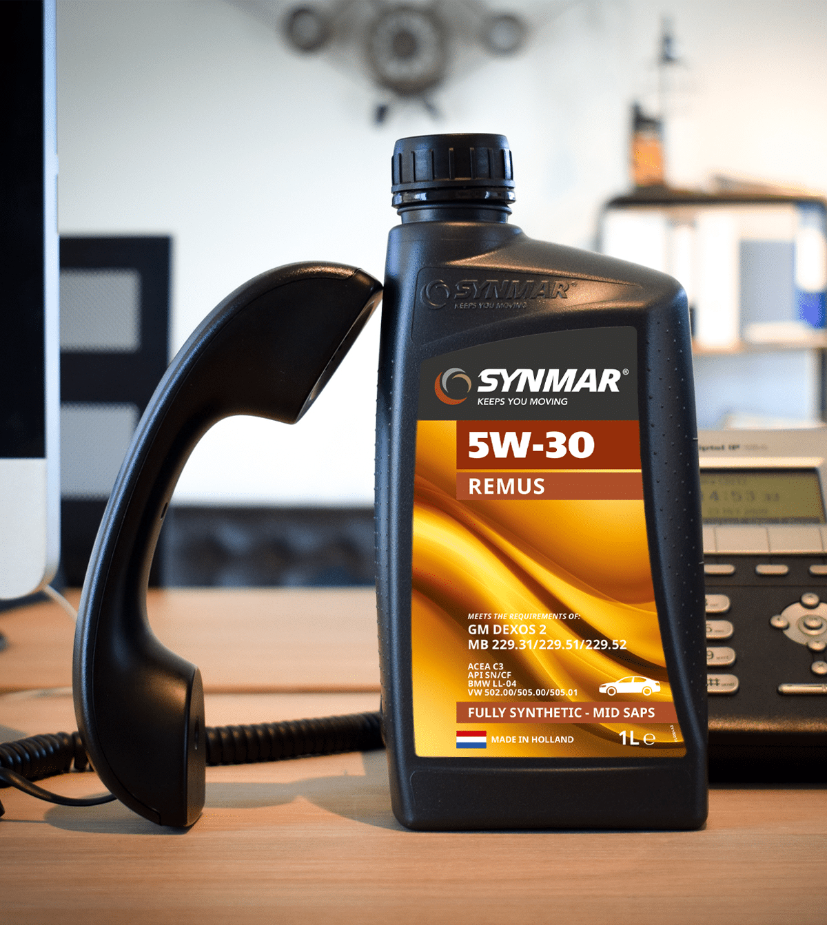 Fles SYNMAR 5W-30 Remus motorolie op een bureau naast een telefoontoestel, met kantoorachtergrond. 