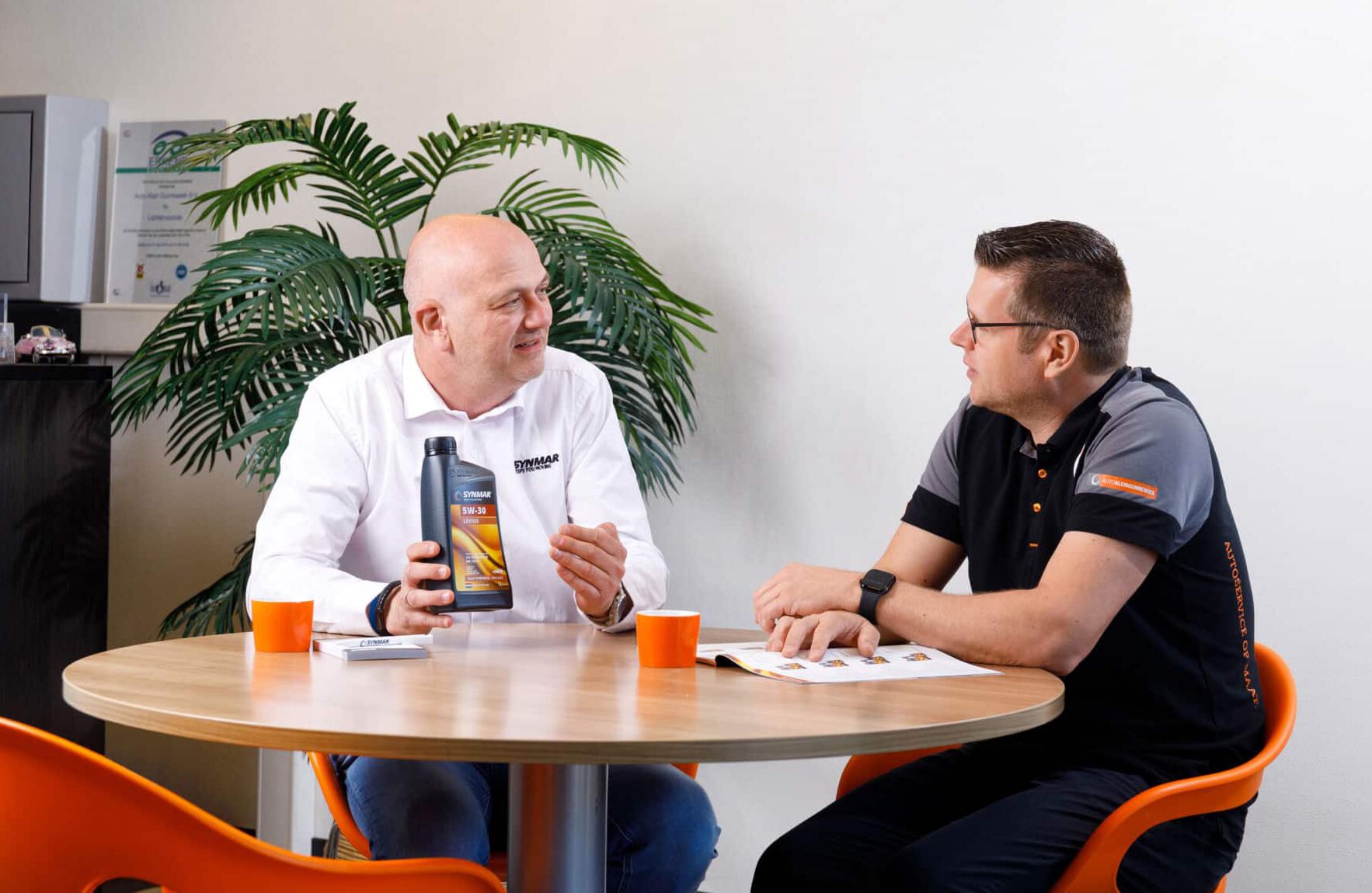 Twee mannen aan een tafel in gesprek over SYNMAR motorolie, met een fles olie op tafel en brochures erbij. 